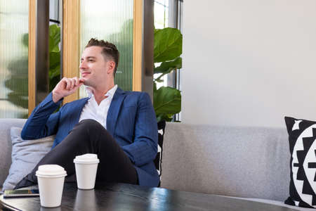 Adult Caucasian businessman dress in smart formal suit sitting inside modern office space drinking coffee while waiting and planning for business meeting. Entrepreneur concept with copy space.の写真素材