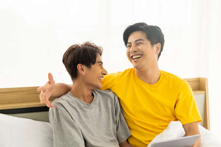 Selective focus at face. Good looking Asian gay homosexual couple hug and smile while using Digital tablet smartphone, showing love expression on the bed. LGBT relationship concept with copy space.の写真素材