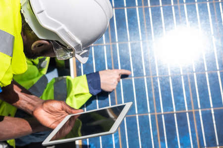 Selective focus at digital tablet. African and Caucasian engineer inspect electrical solar panel and record information into mobile tablet. Alternative energy and industrial concept. Outdoor shot.の写真素材