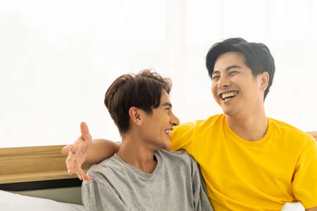 Selective focus at face. Good looking Asian gay homosexual couple hug and smile while using Digital tablet smartphone, showing love expression on the bed. LGBT relationship concept with copy space.の写真素材