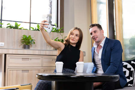 Selective focus at smartphone. Young Caucasian couple dating inside of the coffee shop while women taking selfie photo from mobile phone. Dating activity, modern relationship lifestyle concept.の写真素材