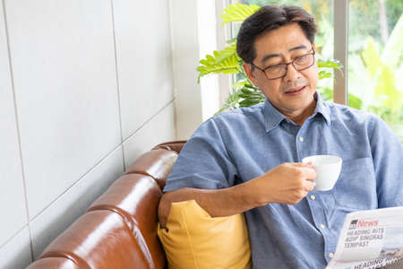 Selective focus at face. Senior Asian man reading newspaper mock up at the sofa couch while drinking coffee in the morning. Happy retirement with relaxation lifestyle and routine.の写真素材
