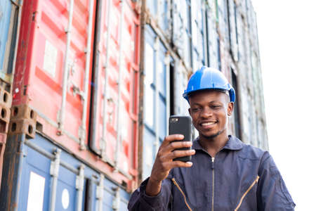 Selective focus at face of Black African man worker wearing safety equipment, talking on smartphone while check and inspection the condition of container. Import and export, logistic freight worker.の写真素材