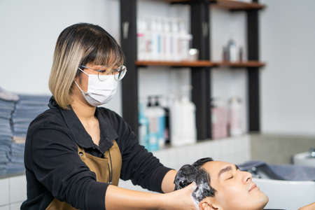 Young Asian women wearing surgical face mask while hair wash with shampoo and massage for her customer. Hair salon studio with hair stylish, beauty and fashion with new normal conceptの写真素材