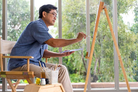 Indoor shot of senior Asian men using brush and oil color to paint on painting canvas. With smile on his face. Happy retirement concept for active senior. With Blurred living room background.の写真素材