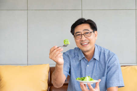 Senior Asian men sitting on the sofa at home while eating green vegetable salad and organic orange juice for breakfast. Healthy eating lifestyle for Elder retirement people concept.の写真素材