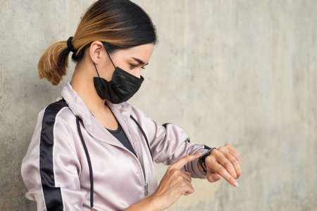 Selective focus at face of young beautiful Asian women wearing surgical face mask using smart watch to track activity before exercise or running at the park in the morning. New normal lifestyle.の写真素材