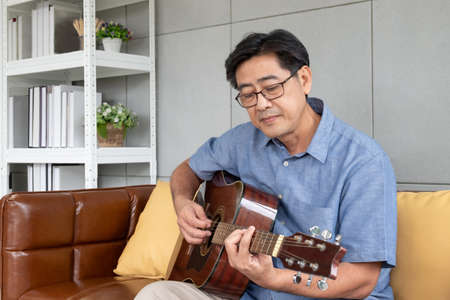 Asian elder man enjoy playing guitar at the sofa inside of well interior decoration house. Active senior lifestyle after retirement with happiness with a good mental health.の写真素材