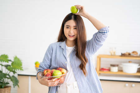 Young Asian women with casual dress in the morning and eat fresh apple fruit for breakfast in the kitchen with different type of vegetable and healthy food. Good nutrition eating human body condition.の写真素材