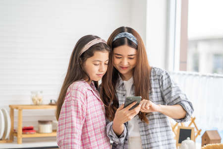 Mixed race girl younger and older sister talk and smile while play and using smartphone inside of the kitchen in the morning while eat breakfast. Family relationship mobile high speed internet networkの写真素材