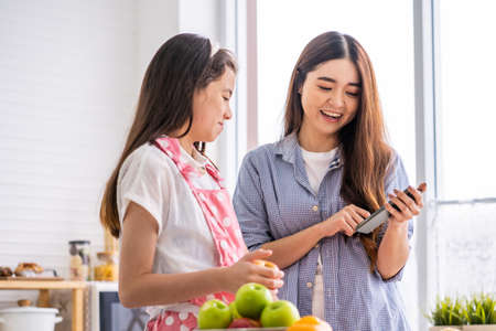Younger and older sister talk and smile while play and using smartphone inside of the kitchen in the morning while eat breakfast. Family relationship mobile high speed internet networkの写真素材