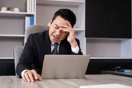 Adult men business person feeling painful from headache while working, online meeting in front of the computer inside of the office area. Office syndrome, over working illness.の写真素材