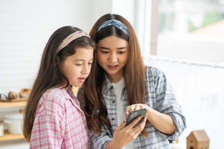Mixed race girl younger and older sister talk and smile while play and using smartphone inside of the kitchen in the morning while eat breakfast. Family relationship mobile high speed internet networkの写真素材