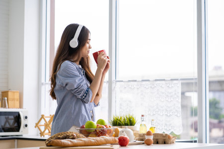 Young Asian women feel relax while drink hot coffee and listen to the music with headphone in the morning inside of kitchen. With breakfast and bread at the background.の写真素材