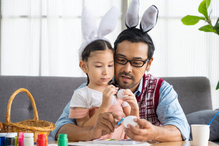 Father and his daughters are painting eggs with colors inside of the living room. Family preparing for Easter. Cute little children girls are wearing bunny ears.の写真素材