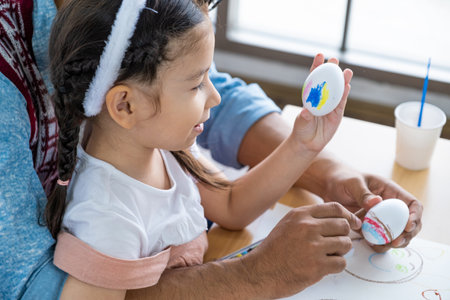 Father and his daughters are painting eggs with colors inside of the living room. Family preparing for Easter. Cute little children girls are wearing bunny ears.の写真素材