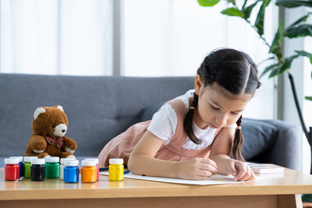Young girl sit on the sofa while using water color and crayon to paint on the paper inside of the living room. Children creativity and learning process, art and education for kidsの写真素材