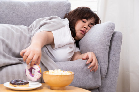 Young Asian oversize women gain weight while eating donut, bread and snack while lay down and sleep at sofa. Junk food that can cause obesity and unhealthy for human body.の写真素材