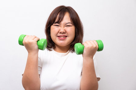 Young Asian chubby woman hold dumbbell for exercise and weight training. Fighting while diet during weight loss program with positive emotion. Portrait shot on white background.の写真素材