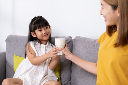 Young Asian mother hand over glass of fresh milk to her daughter inside of living room. Children who encourage to drink milk for good protein and nutrition.の写真素材