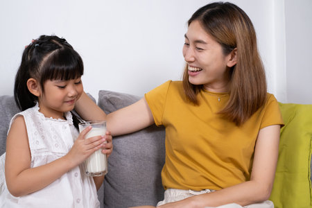Young Asian mother hand over glass of fresh milk to her daughter inside of living room. Children who encourage to drink milk for good protein and nutrition.の写真素材