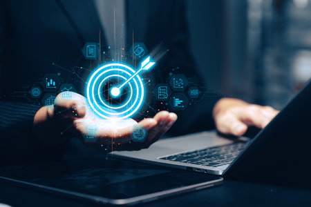 Businessman holding a glowing blue target with arrow in his palm above a laptop, surrounded by holographic business icons representing goals, analytics, strategy, and online presence. This conceptual image embodies business target achievement, success, digital marketing strategy, online business goals, data-driven decision making, technological focus, and aiming for results in the modern digital landscape.の写真素材