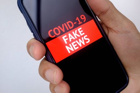 A hand holding a mobile phone with the words Covid-19 Fake News flashing on the screen. white...