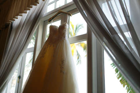 A beautiful white wedding dress hanging beside a windowの写真素材