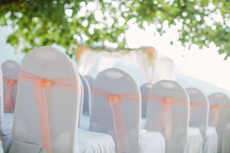 View of chairs in white covers with pink ribbons during a beach weddingの写真素材