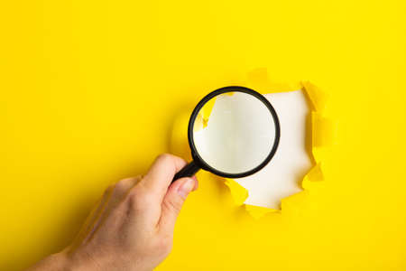 A human hand holds a magnifying glass through a torn hole in a yellow paper.