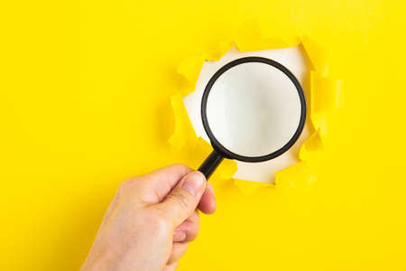 A human hand holds a magnifying glass through a torn hole in a yellow paper.の写真素材