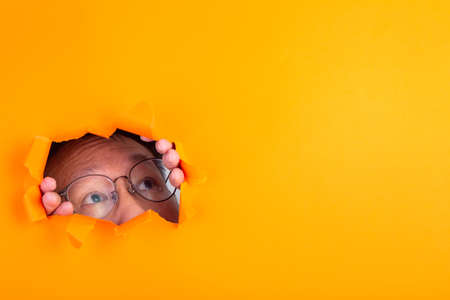 The man with spectacle peeks through a cut hole in the orange paper background.の写真素材
