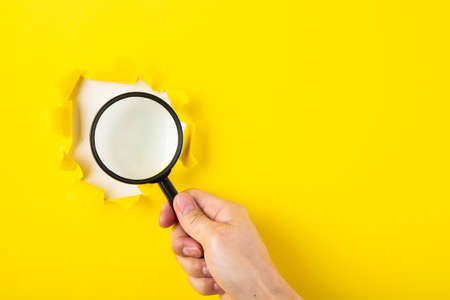 A human hand holds a magnifying glass through a torn hole in a yellow paper.の写真素材