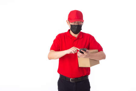 young Asian delivery man in red uniform with  face mask, carrry cardboard box in hands isolated on white background. Delivery conceptの写真素材