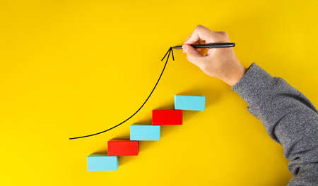 Male hand drawing an upward pointing arrow on top of growing graph made of wooden blocks over yellow backgroundの写真素材