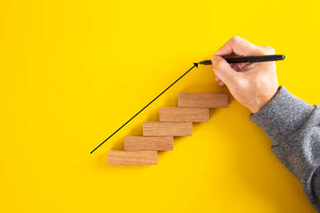 Man hand drawing an upward pointing arrow on top of growing graph made of wooden block. Over yellow...