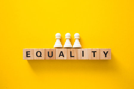 Three white pawns standing on wooden cubes spelling equality on yellow backgroundの写真素材