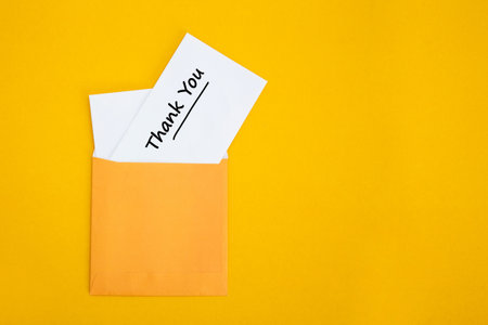 White thank you note partially emerging from a yellow envelope on a solid backgroundの写真素材