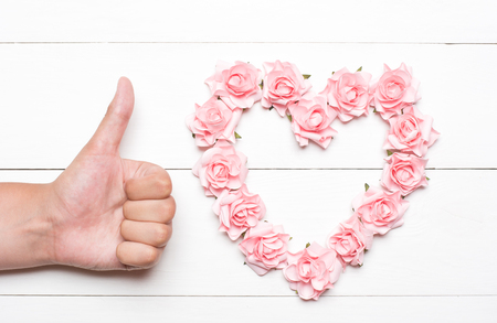 Heart made of pink roses and young hand thumbs up on white wooden table conceptの写真素材