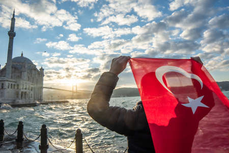 Man hold Turkish flag. Sunrise near Bosphorus bridge (aka: 15 July Martyrs Bridge Turkish: 15 Temmuz Sehitler Koprusu) and Imperial Mosque of Sultan Abdulmecid in Ortakoy, Istanbul, Turkey. Patriotismの写真素材