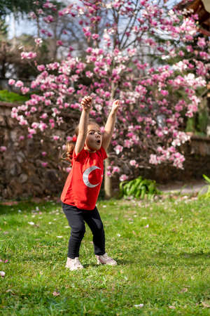 A little girl in a red T-shirt and leggings stands in the garden with a blooming magnolia tree.の写真素材