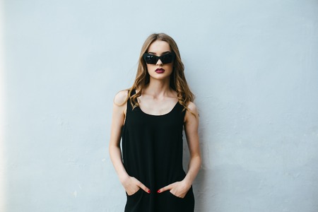 Attractive fashion woman in black dress with sunglasses posing near white wallの写真素材