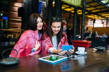 Two young and beautiful girl sitting at the table and doing selfie in the cafe