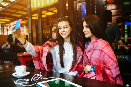Two young and beautiful girl sitting at the table and doing selfie in the cafe