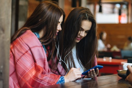 Two young and beautiful girl sitting at the table listening to music with a smartphoneの写真素材