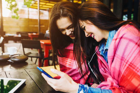 Two young and beautiful girl sitting at the table listening to music with a smartphoneの写真素材