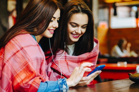 Two young and beautiful girl sitting at the table listening to music with a smartphoneの写真素材