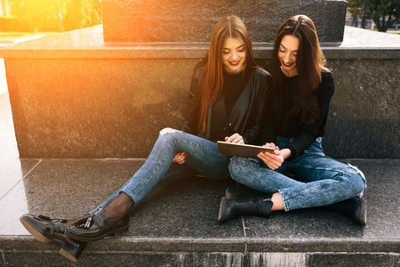 Two young adult women sitting in the park and looking something on a tabletの写真素材