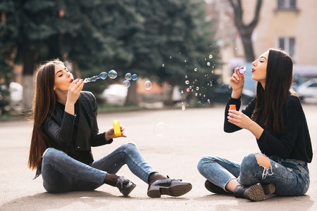Two young adult girls sitting in a park and blow bubblesの写真素材