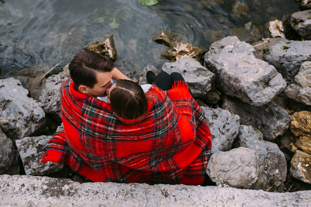 beautiful young couple sitting on a stone embankment, wrapped in a blanketの写真素材